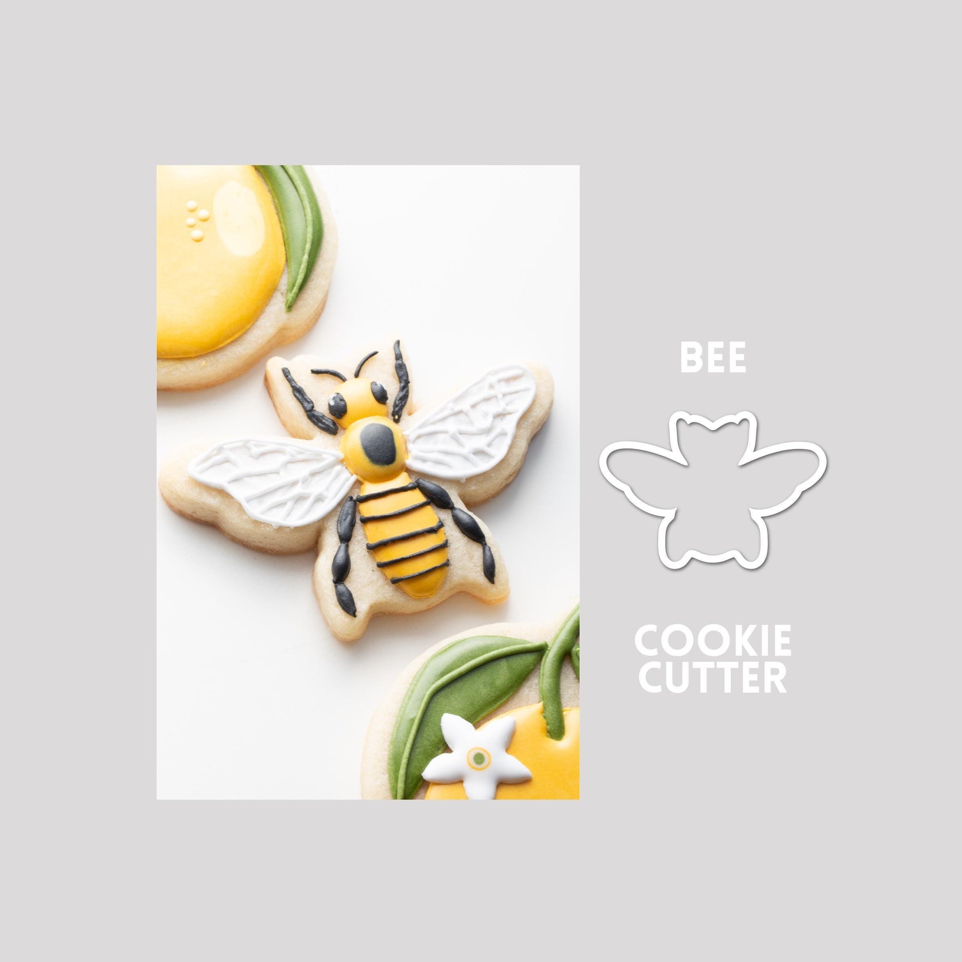 Decorated bee sugar cookie next to a bee cookie cutter outline on a gray background.