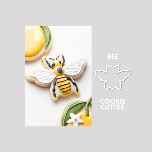 Decorated bee sugar cookie next to a bee cookie cutter outline on a gray background.