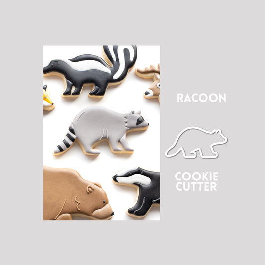 Decorated raccoon sugar cookie next to a raccoon cookie cutter outline on a gray background.