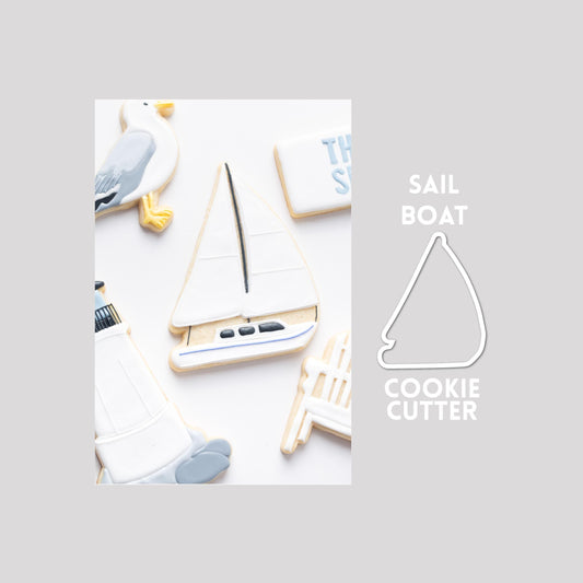 Decorated sailboat sugar cookie next to a sailboat cookie cutter outline on a gray background.