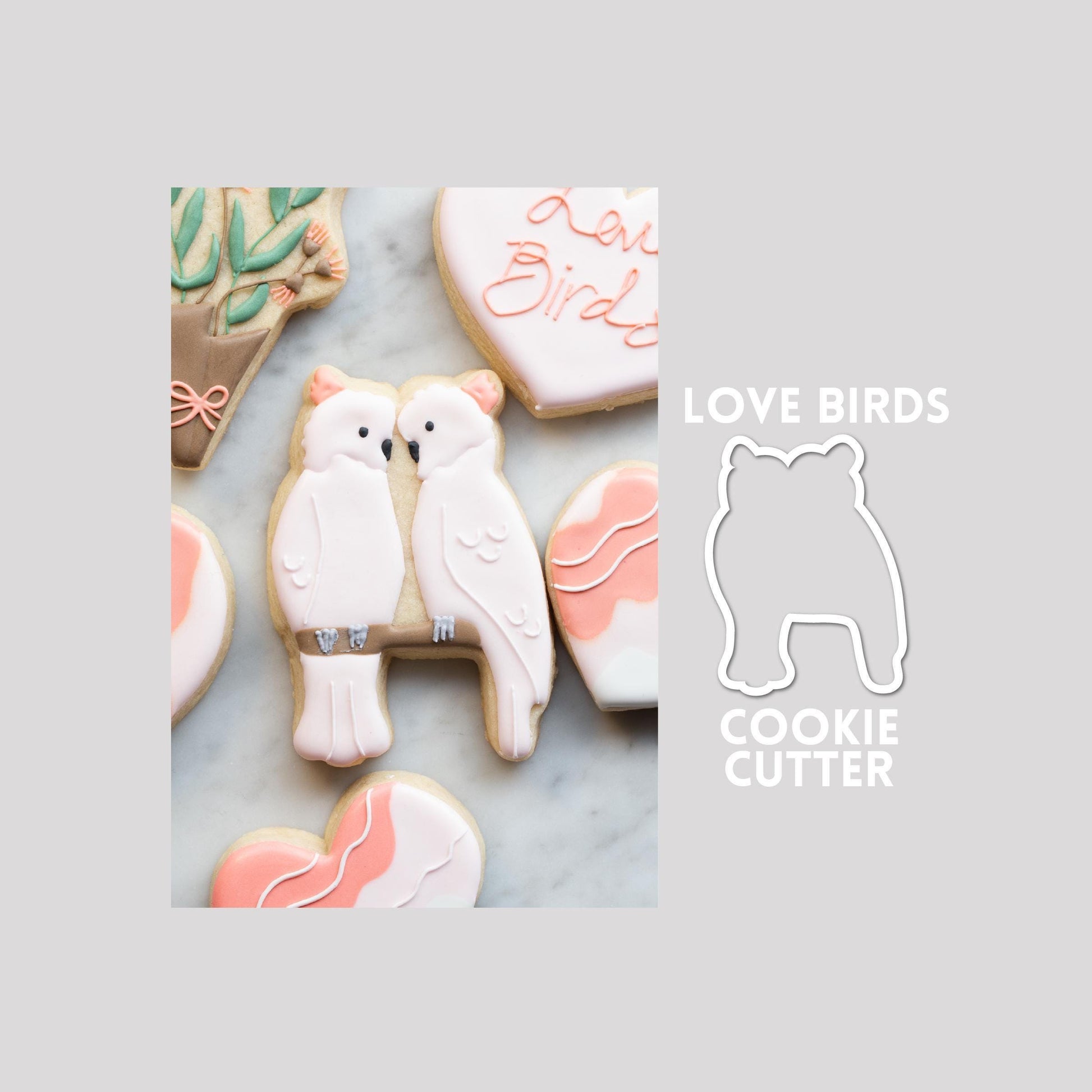 Decorated love birds sugar cookie next to a love birds cookie cutter outline on a gray background.