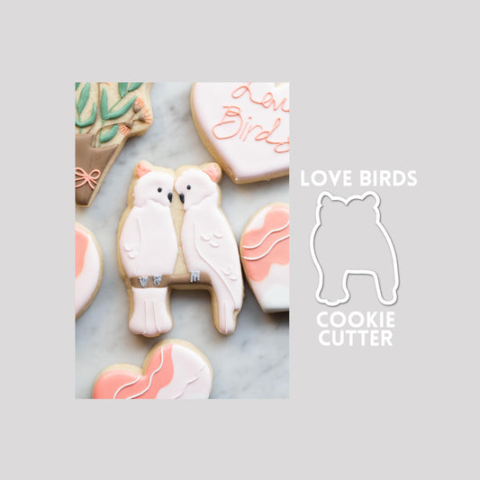Decorated love birds sugar cookie next to a love birds cookie cutter outline on a gray background.