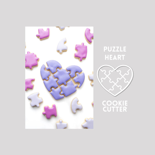 Decorated puzzle heart sugar cookie with puzzle pieces next to a puzzle heart cookie cutter outline on a gray background.