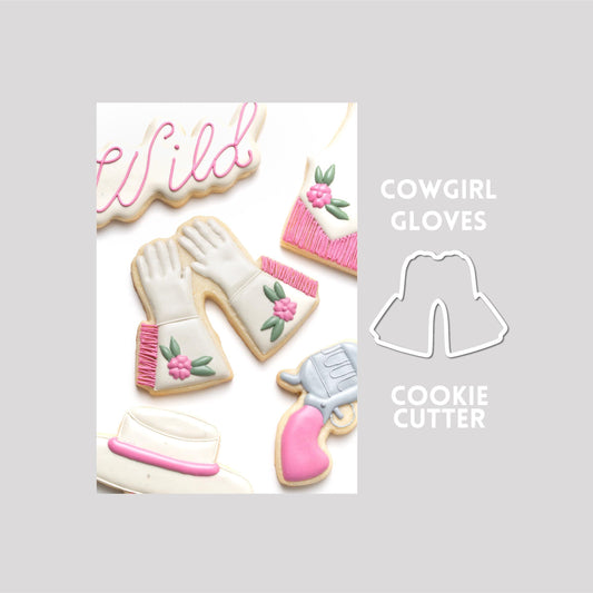 Decorated cowgirl gloves sugar cookie next to a cowgirl gloves cookie cutter outline on a gray background.