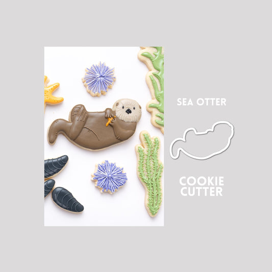 Decorated otter sugar cookie next to an otter cookie cutter outline on a gray background.