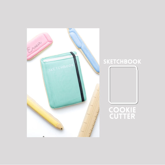 Decorated sketchbook sugar cookie next to a sketchbook cookie cutter outline on a gray background.