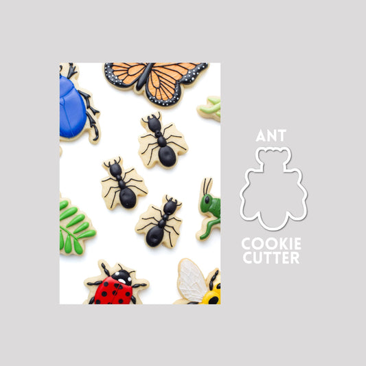 Decorated ant sugar cookie next to a ant cookie cutter outline on a gray background.