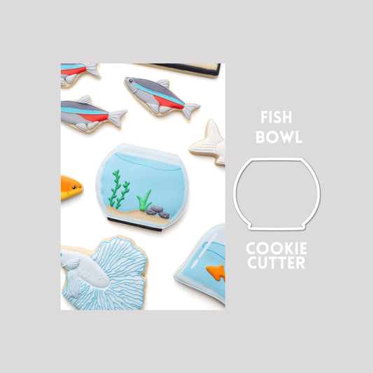 Decorated fish bowl sugar cookie next to a fish bowl cookie cutter outline on a gray background.