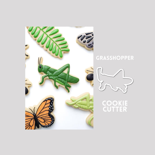 Decorated grasshopper sugar cookie next to a grasshopper cookie cutter outline on a gray background.