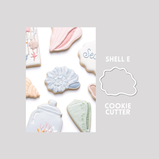 Decorated seashell sugar cookie next to a seashell cookie cutter outline on a gray background.