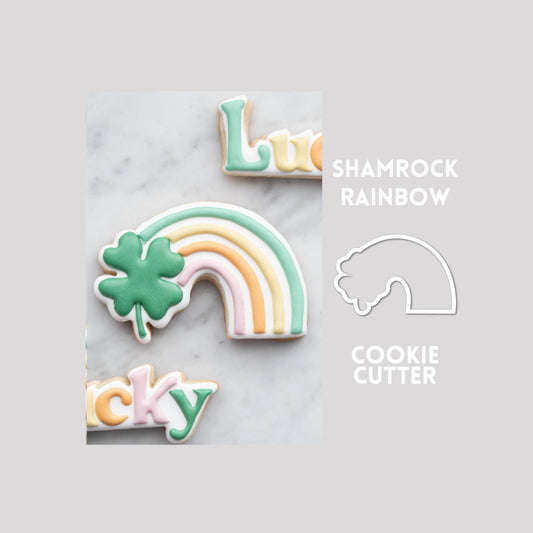 Decorated shamrock rainbow sugar cookie next to a shamrock rainbow cookie cutter outline on a gray background.