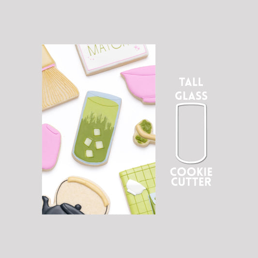 Decorated tall glass with matcha tea and ice sugar cookie next to a tall glass with matcha tea and ice cookie cutter outline on a gray background.