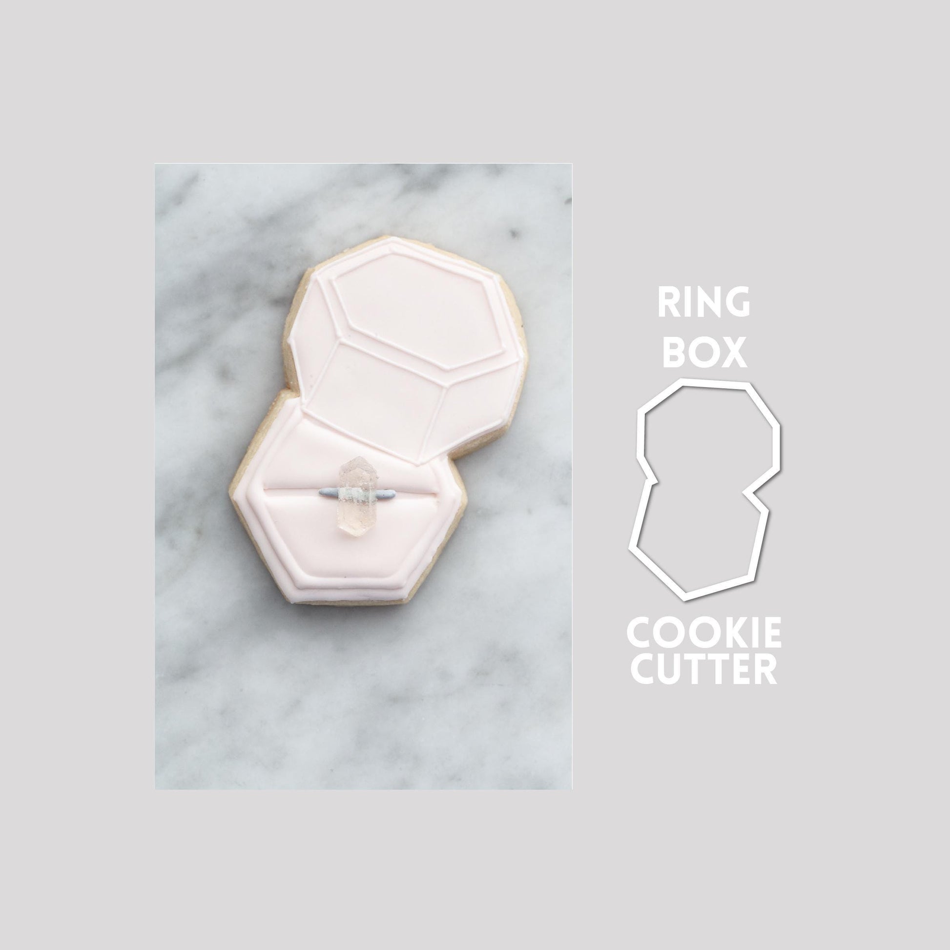 Decorated wedding box sugar cookie next to a wedding box cookie cutter outline on a gray background.