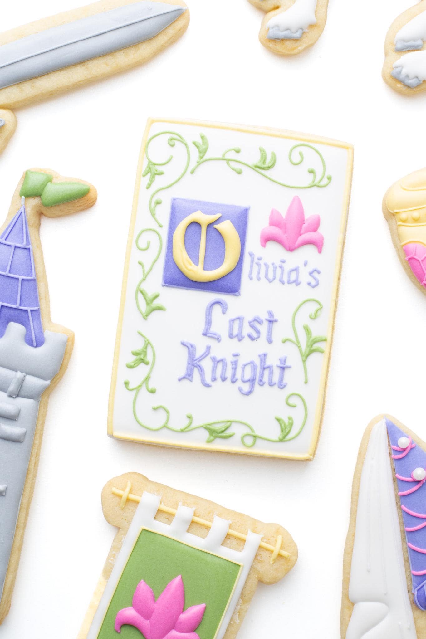 Medieval manuscript titled “Olivia’s Last Knight” decorated sugar cookie next to a medieval hanging banner, goblet, knight's sword, castle tower and princess hat. 