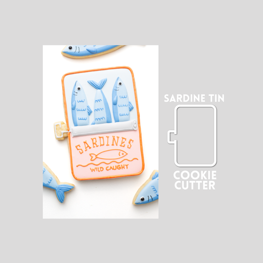 Decorated open tin or can of sardine fish sugar cookie next to a sardine tin or can cookie cutter outline on a gray background.