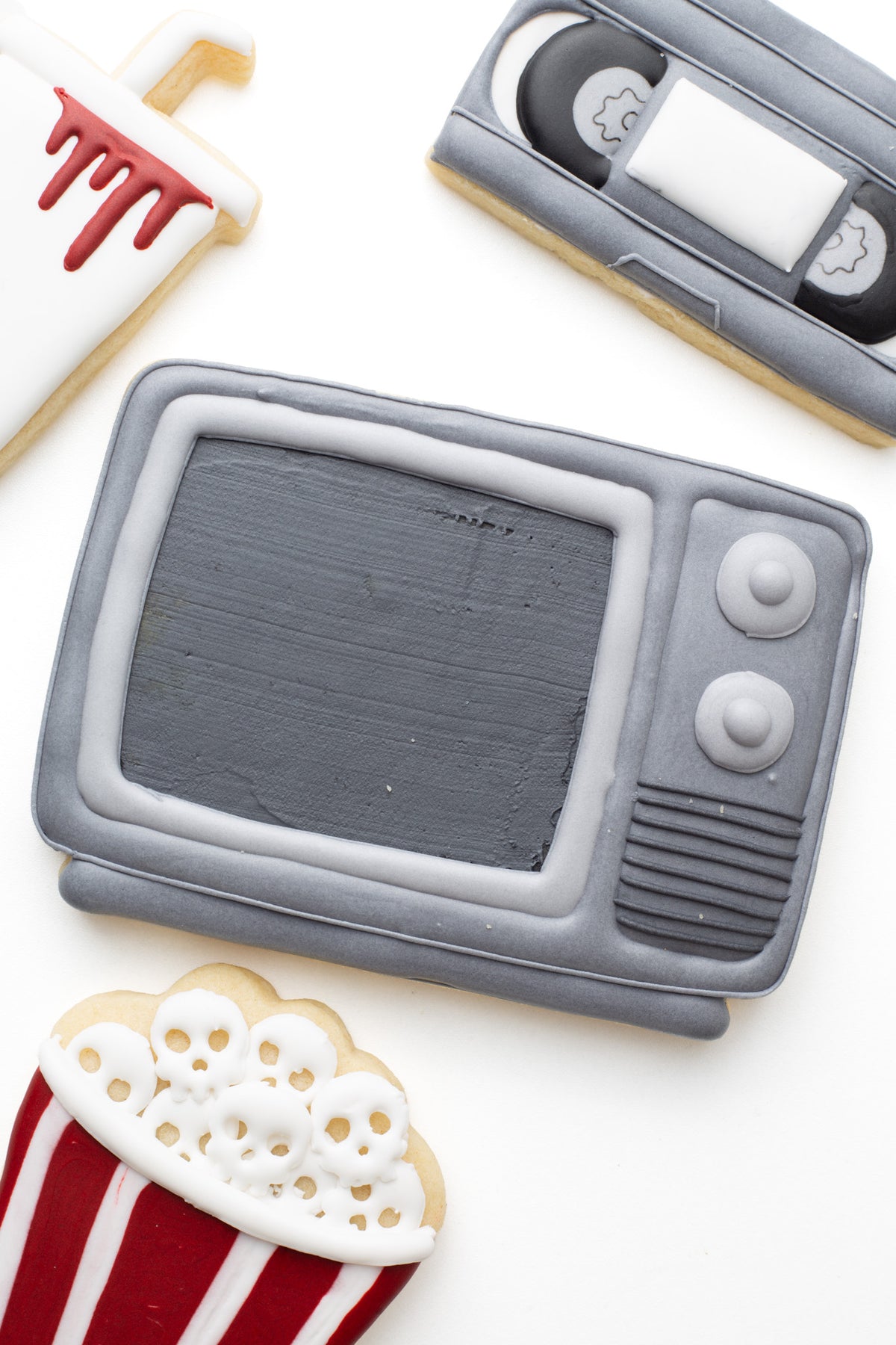 A television decorated sugar cookie next to a video tape, and popcorn tub and cup with blood and skulls in a scary movie halloween theme. 