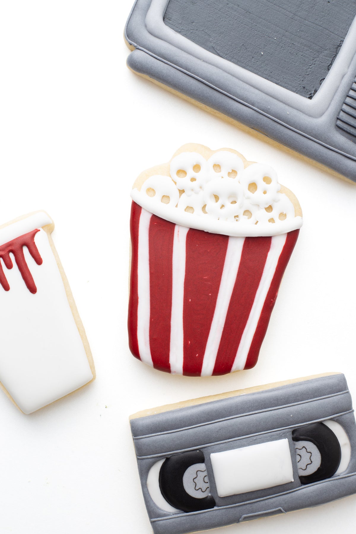 A popcorn tub decorated sugar cookie with blood and skulls next to a television, video tape, and cup with blood  in a scary movie halloween theme. 