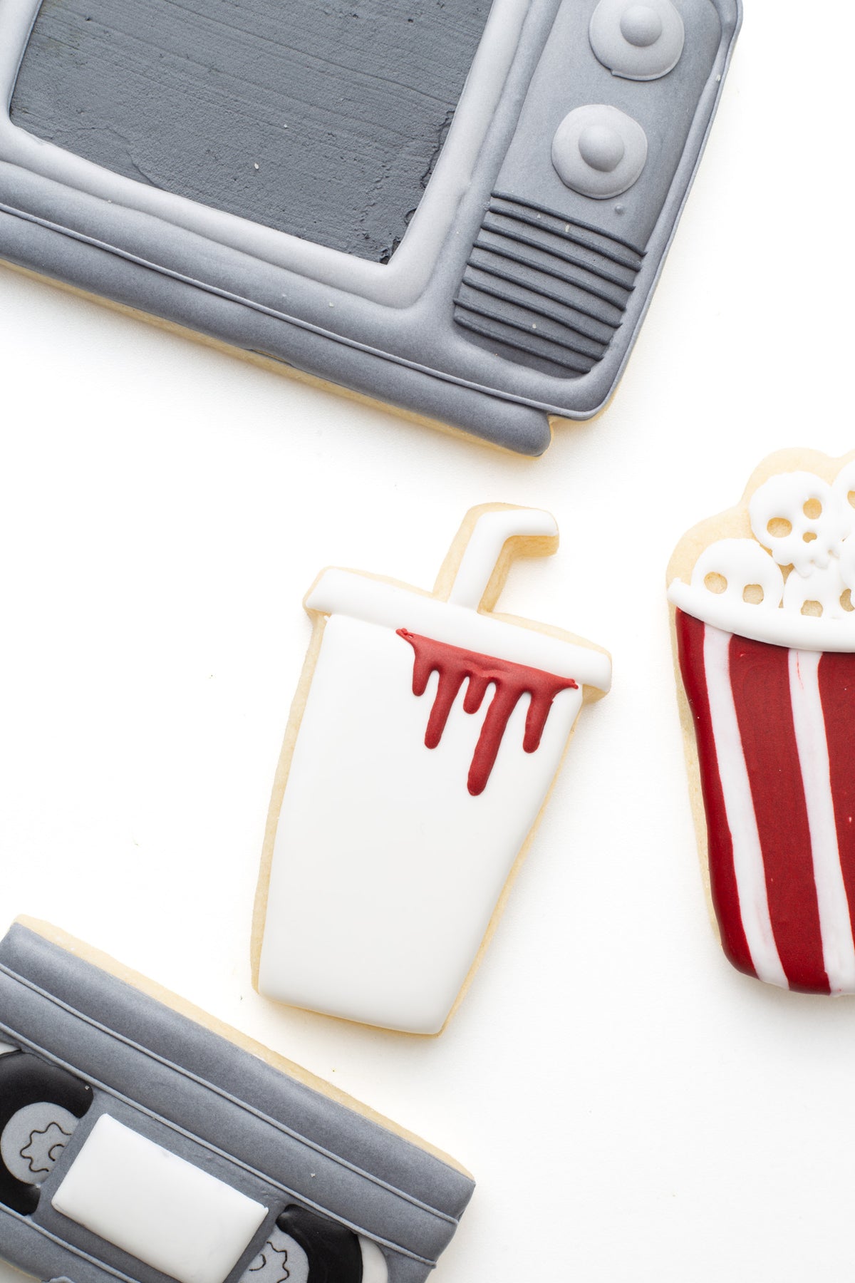 A  fountain cup decorated sugar cookie with blood next to a television, video tape, and popcorn tub with skulls  in a scary movie halloween theme. 