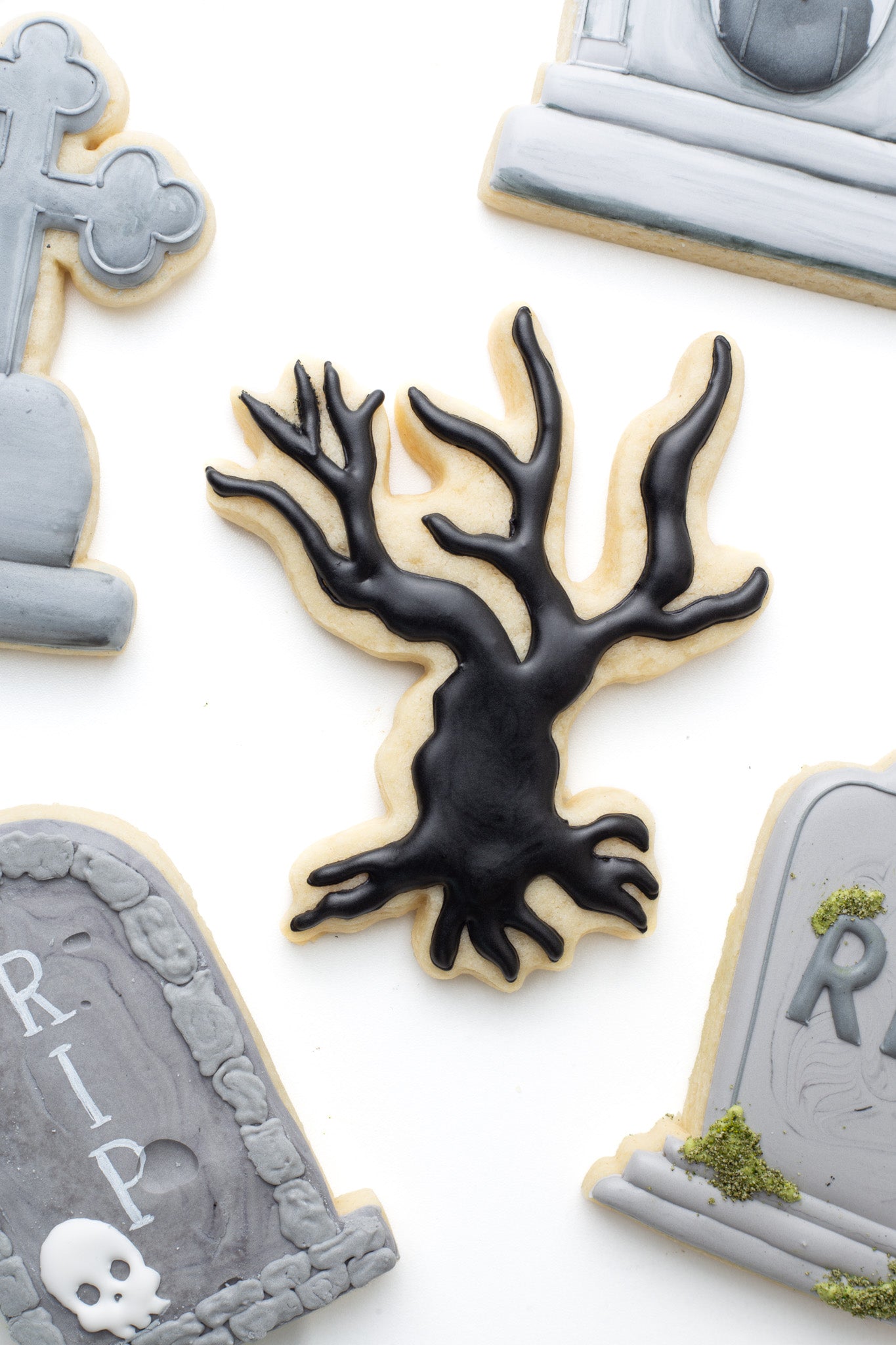 A spooky tree decorated sugar cookie next to gravestones decorated sugar cookies.
