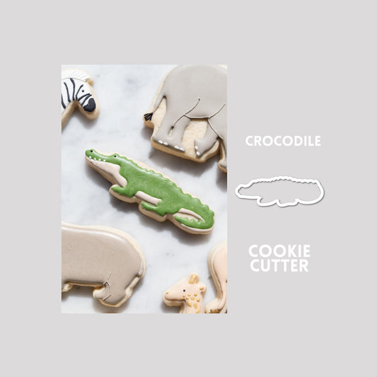 Decorated crocodile sugar cookie next to a crocodile cookie cutter outline on a gray background.