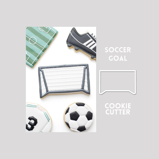 Decorated soccer goal sugar cookie next to a soccer goal cookie cutter outline on a gray background.