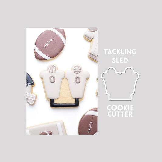 Decorated tackling sled sugar cookie next to a tackling sled cookie cutter outline on a gray background.