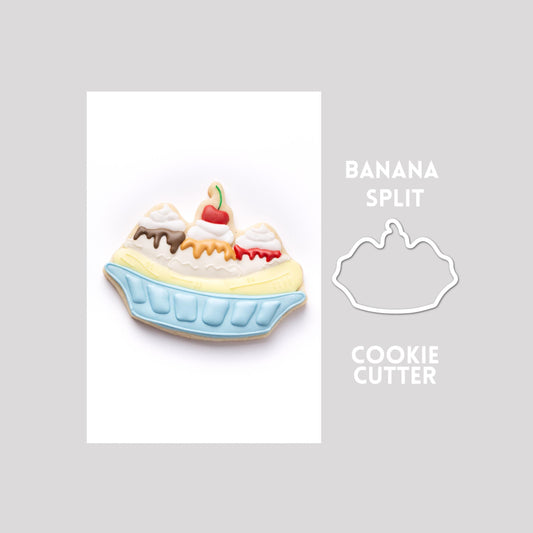 Decorated banana split sugar cookie next to a banana split cookie cutter outline on a gray background.