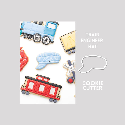 Decorated train engineer hat sugar cookie next to a train engineer hat cookie cutter outline on a gray background.