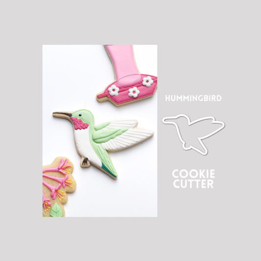 Decorated hummingbird sugar cookie next to a hummingbird cookie cutter outline on a gray background.