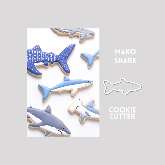 Decorated mako shark sugar cookie next to a mako shark cookie cutter outline on a gray background.