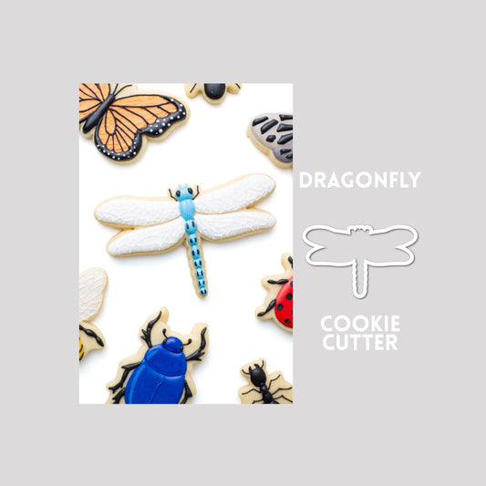 Decorated dragonfly sugar cookie next to a dragonfly cookie cutter outline on a gray background.
