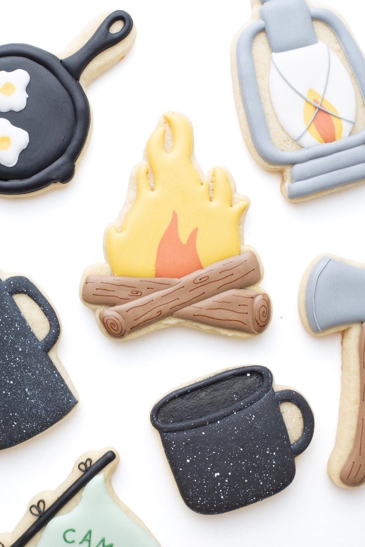 Campfire decorated sugar cookie next to a lantern, axe, lantern, camp mug and kettle, cast iron pan, and camp pennant flag decorated sugar cookies. 