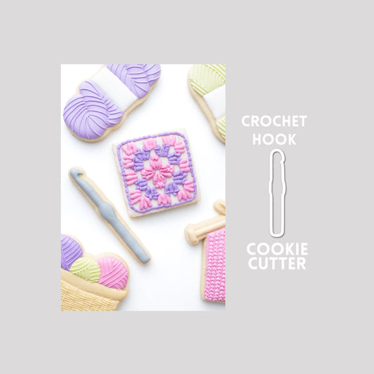 Decorated crochet hook sugar cookie next to a crochet hook cookie cutter outline on a gray background.