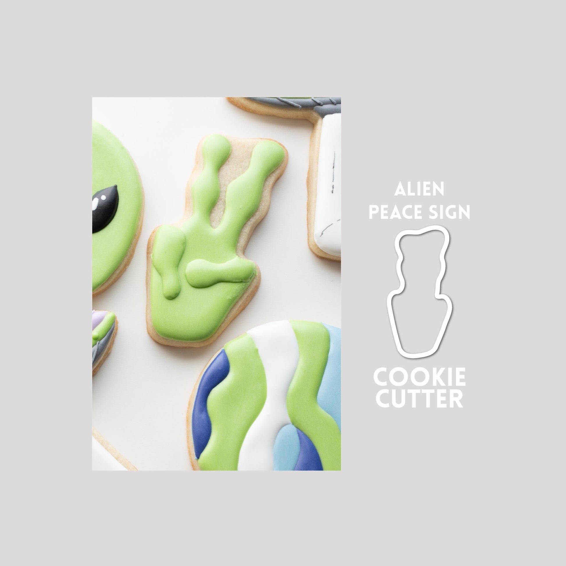 Decorated alien peace sign sugar cookie next to a alien peace sign cookie cutter outline on a gray background.