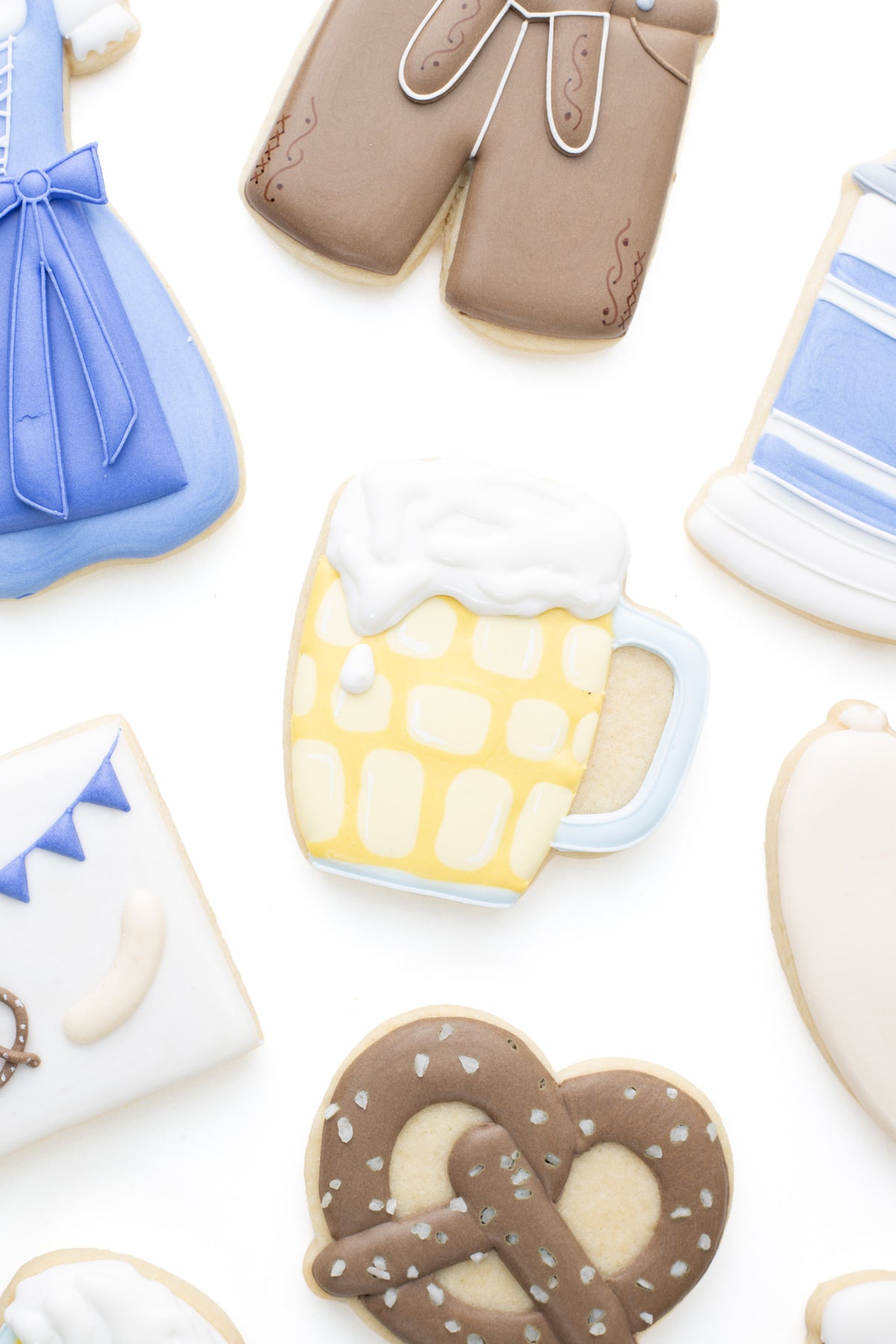 Oktoberfest themed cookies. A dimple mug decorated sugar cookie next to a lederhosen,  beer stein, pretzel and bratwurst decorated sugar cookie.