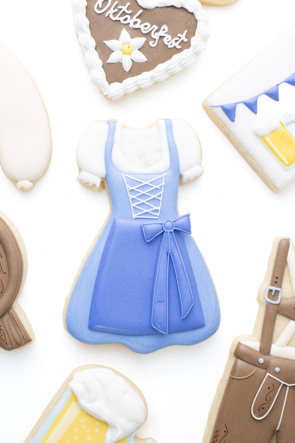 Oktoberfest themed cookies. A dirndl dress decorated sugar cookie next to lederhosen, beer mug, beer keg, bratwurst, and lebkuchenherzen decorated sugar cookies.