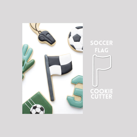Decorated soccer flag sugar cookie next to a soccer flag cookie cutter outline on a gray background.