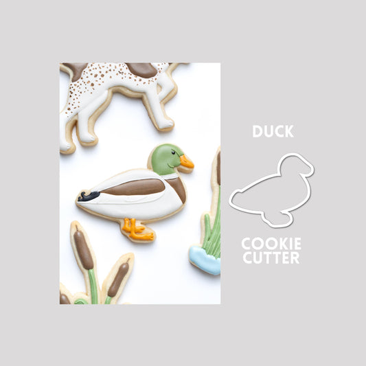 Decorated duck sugar cookie next to a duck cookie cutter outline on a gray background.