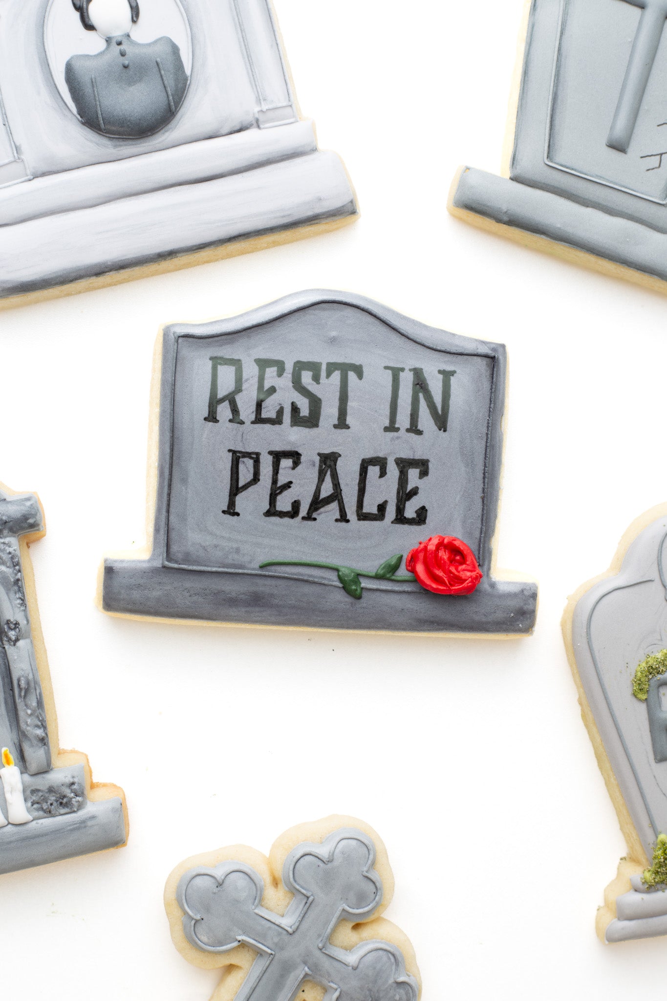 A gravestone decorated sugar cookie featuring the words “REST IN PEACE,” accented with a small red rose, displayed alongside other spooky gravestone-themed decorated sugar cookies.