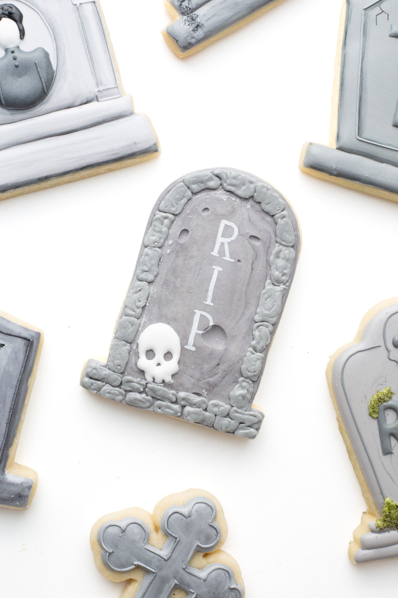 Decorated gravestone decorated sugar cookie with a skull and stones and 'RIP' lettering surrounded by other gravestone decorated sugar cookies. 