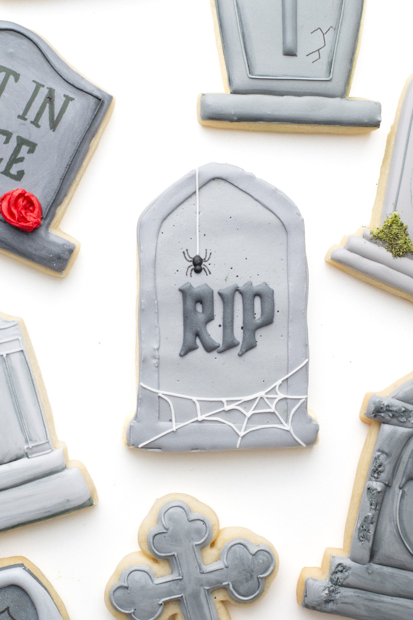 A gravestone decorated sugar cookie with the words 'RIP' , spider and spider web surrounded by other gravestone decorated sugar cookies. 