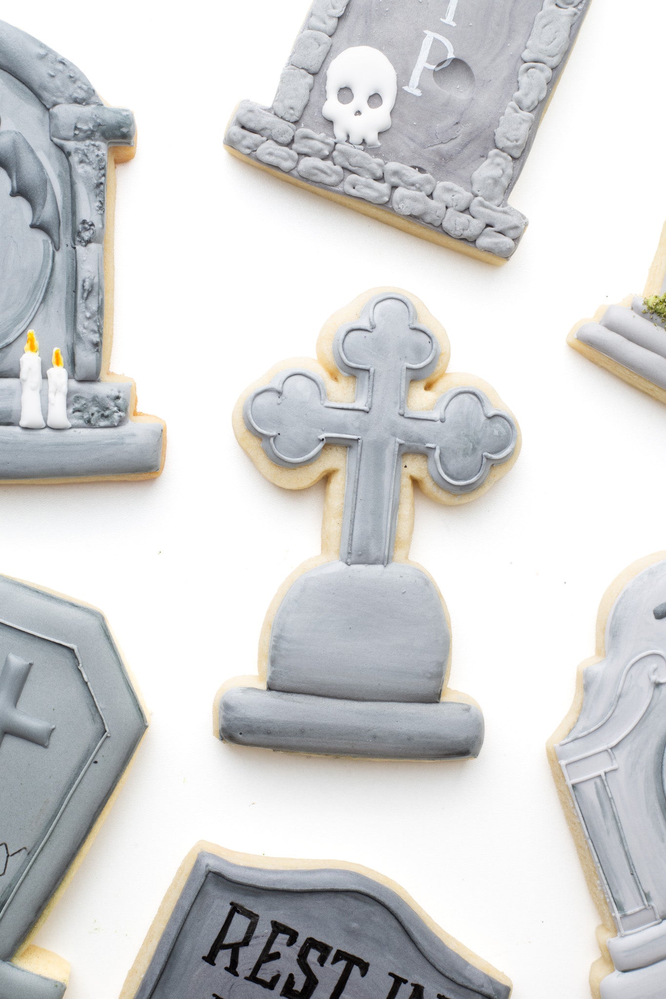 A gravestone decorated sugar cookie shaped like a cross decorated by other gravestone decorated sugar cookies. 