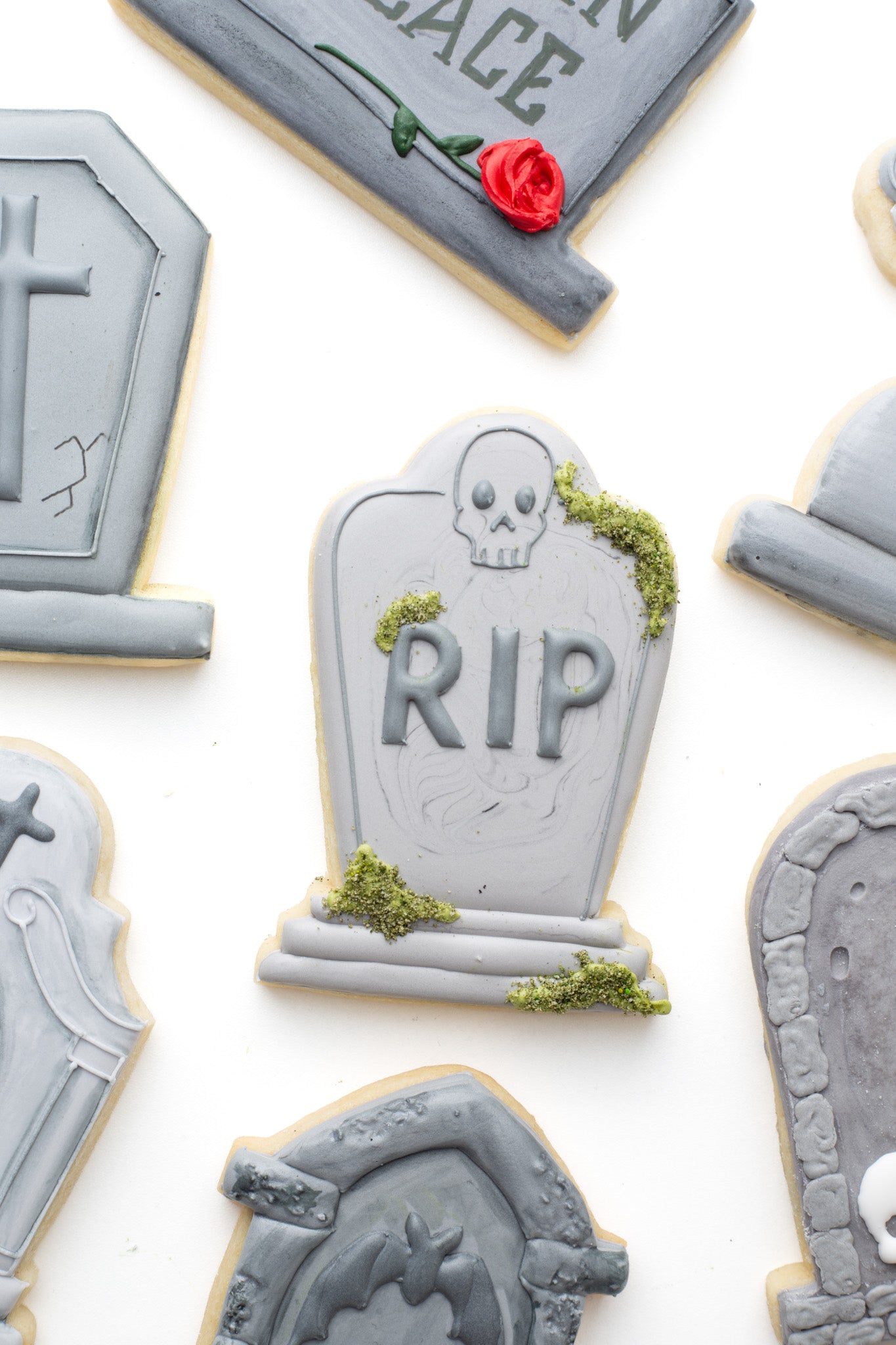 Gravestone decorated sugar cookie with a skull, green moss accents surrounded by gravestone decorated sugar cookies. 