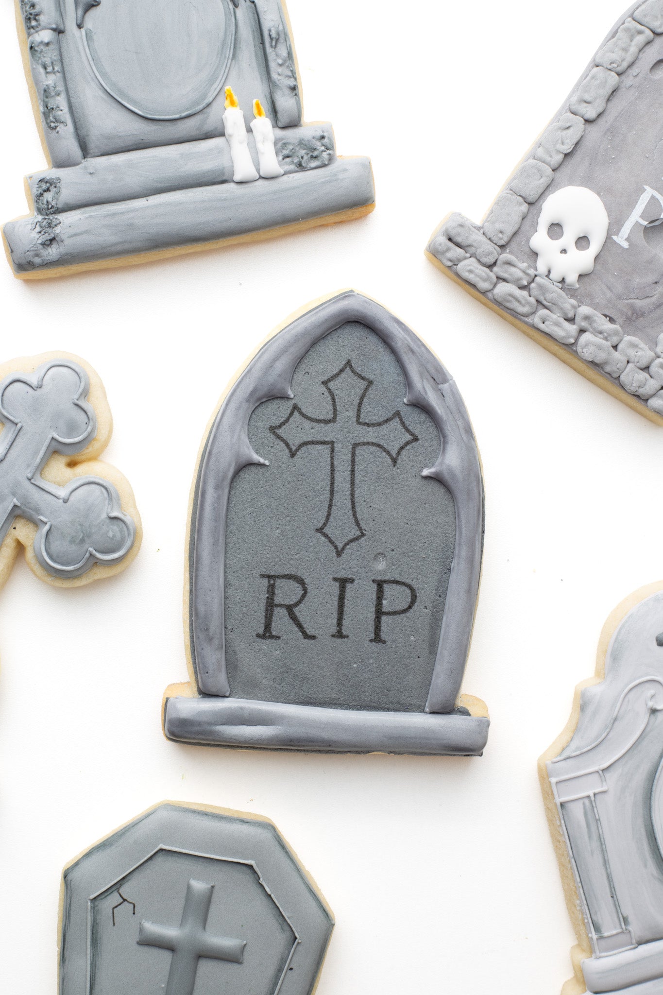 A gravestone decorated sugar cookie with a gothic cross and words'RIP' surrounded by gravestone decorated sugar cookies. 