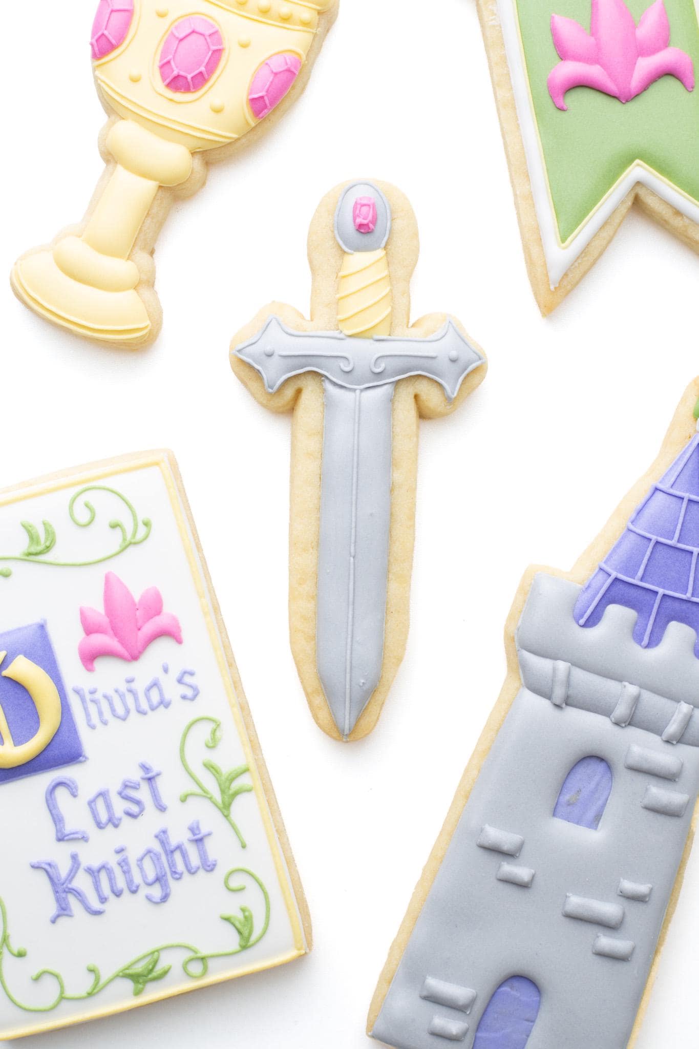 Knight’s sword decorated sugar cookie next to a castle, medieval banner, goblet, and a medieval manuscript titled “Olivia’s Last Knight” as a set of fantasy themed cookies. 