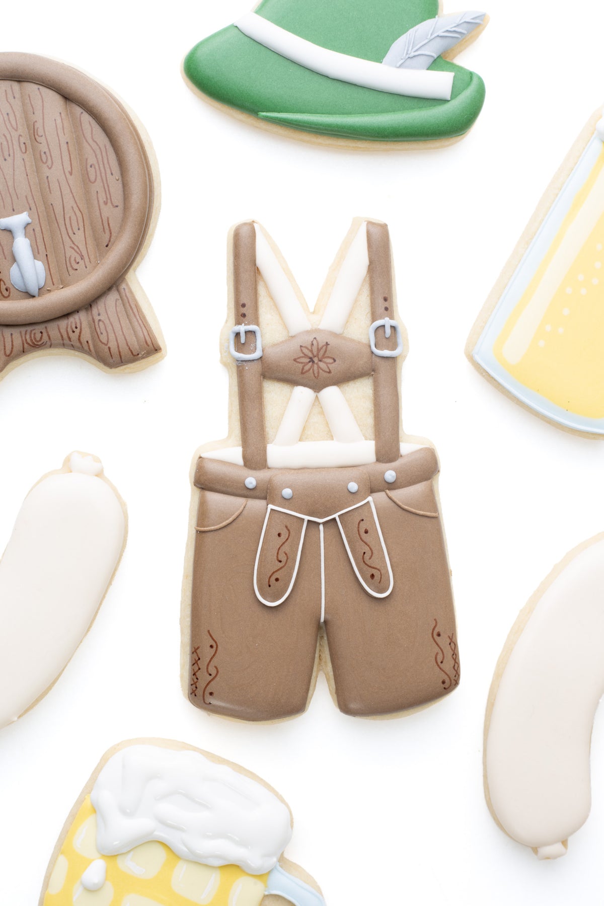 Oktoberfest themed cookies. A lederhosen decorated sugar cookie next to a keg, beer, beer stein, and bratwurst decorated sugar cookies.