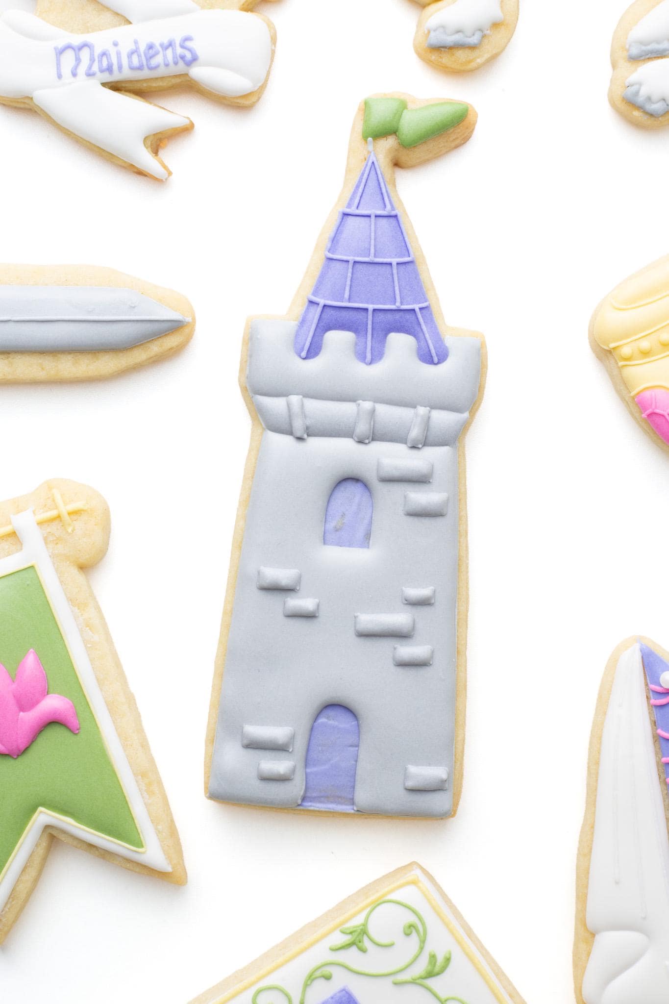 Castle tower decorated sugar cookie next to a medieval banner, goblet, and knight’s sword as a set of fantasy themed cookies. 