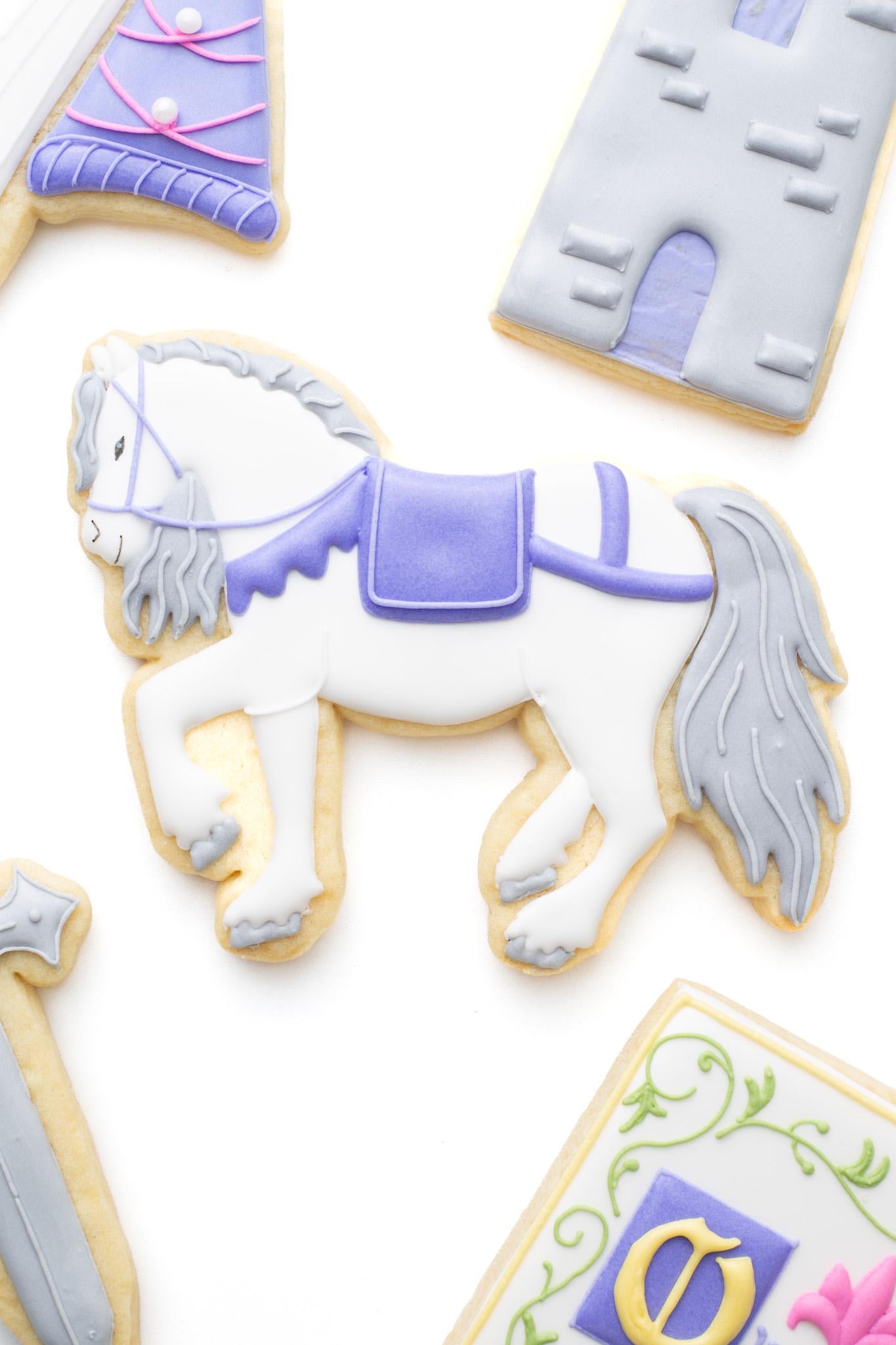 Medieval horse decorated sugar cookie next to a castle tower, princess hat, knight's sword and medieval manuscript  titled “Olivia’s Last Knight”.