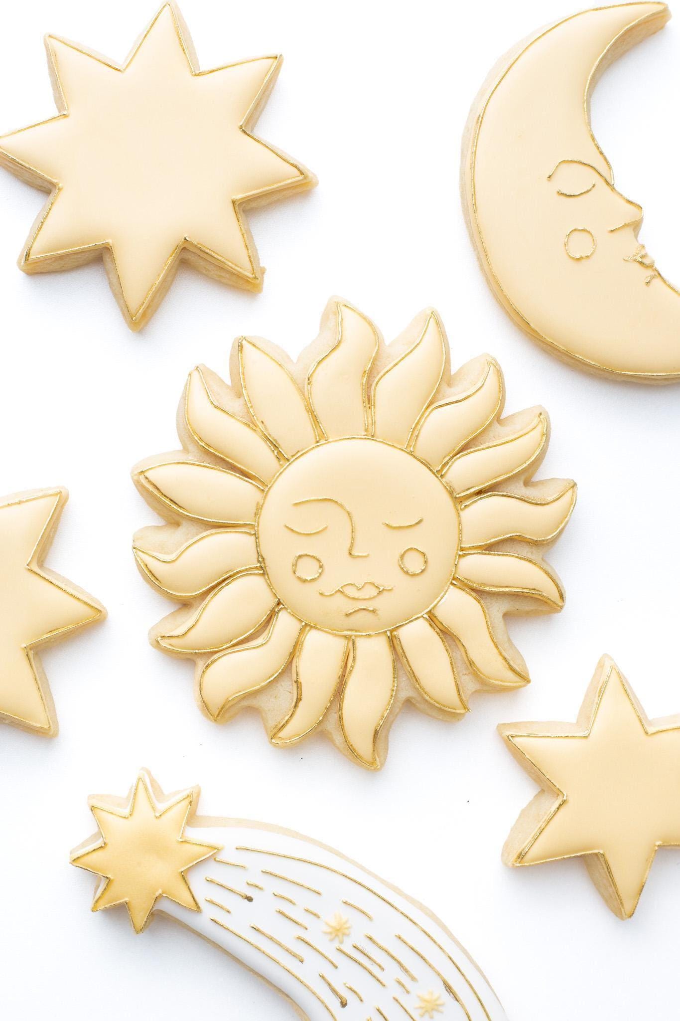 A celestial sun with a face decorated sugar cookie next to a shooting star, celestial crescent moon with a face and a 6 point,7 point, and 8 point star.