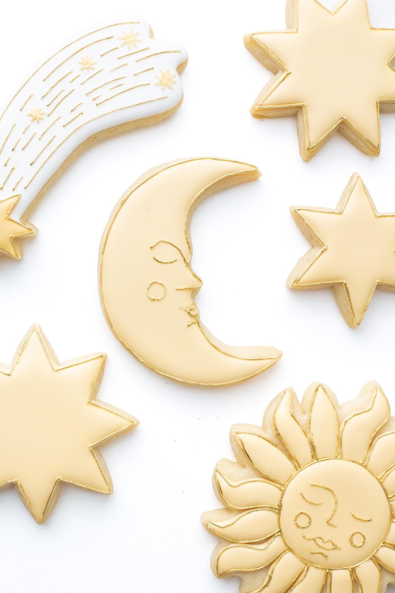 A celestial crescent moon with a face decorated sugar cookie next to a shooting star, celestial sun with a face and a 6 point,7 point, and 8 point star.